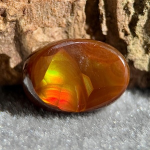 Op de afbeelding: Een gepolijste, ovaalvormige edelsteen met een vurig kleurenspectrum. De steen gaat over van diep rood en oranje naar levendig geel en groen. Het oppervlak van de edelsteen is glad en reflecterend.