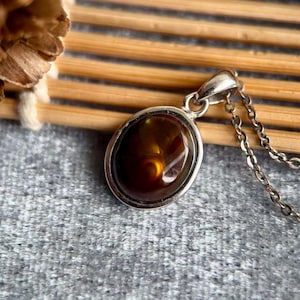 Op de afbeelding: Ketting met ovale hanger en zilveren ketting. De hanger heeft een gepolijste, bruine edelsteen met een gouden binnenkant. De ketting wordt getoond op een grijze ondergrond met een houten achtergrond.