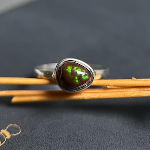 May include: A silver ring featuring a teardrop-shaped dark brown stone with vibrant green iridescent flashes. The stone is set in a smooth metal bezel, and the band is polished silver. The ring is positioned horizontally on a thin wooden stick.