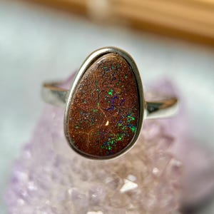 May include: A silver ring featuring a teardrop-shaped boulder opal gemstone. The stone has a dark brown, textured surface with vibrant, iridescent flecks of green and blue light reflecting across its face, set in a smooth, polished silver bezel.