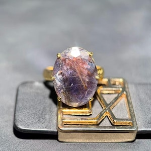 May include: A gold ring featuring a large, oval-shaped faceted gemstone with a translucent appearance, displaying deep purple and reddish-brown inclusions throughout. The stone is held in a gold prong setting atop a geometric gold band structure.