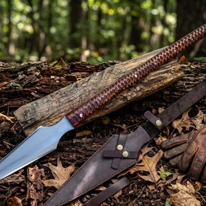 May include: A long spear with a polished silver metal blade and a textured dark brown wooden handle featuring silver rivets. Beside the spear lies a dark brown leather sheath with brass snap closures and a pair of brown leather work gloves.
