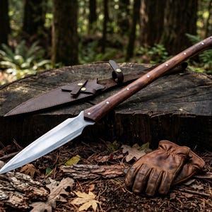 May include: A long spear with a sharp, pointed metal blade and a dark brown wooden handle secured with small metal rivets. Beside the spear lies a matching brown leather sheath with metal snap closures and a pair of tan leather work gloves.