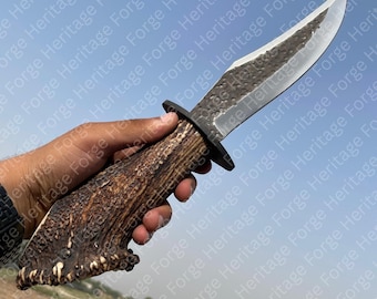 May include: A handcrafted hunting knife with a textured blade and a handle made from a brown antler. The knife has a black guard and a sharp, curved blade. The blade is approximately 20 cm long.