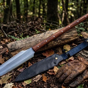 May include: A long spear with a polished metal blade and a dark brown wooden handle secured by silver rivets. Beside the spear lies a black leather sheath with a brass snap closure and a pair of worn brown leather work gloves on the forest floor.