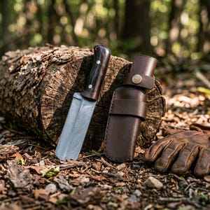 May include: A fixed-blade knife with a dark wood handle and a silver metal blade featuring a subtle pattern, accompanied by a brown leather sheath with a metal snap closure and a pair of brown leather work gloves resting on the ground.
