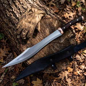 May include: A fixed-blade knife with a curved silver steel blade, a dark wood handle with three metal rivets, and a copper bolster. The knife is accompanied by a black leather sheath and a pair of tan leather work gloves resting on a log.