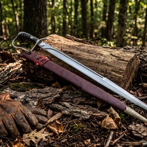 May include: A silver-toned metal sword with a black wrapped handle and a curved hand guard, accompanied by a dark brown leather scabbard featuring a decorative metal tip and a pair of tan leather work gloves resting on the forest floor.