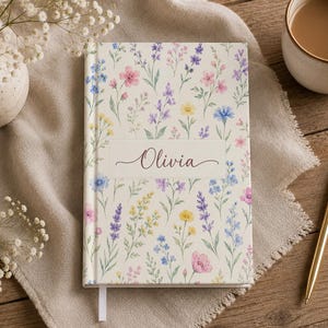 May include: A hardcover journal with a cream-colored cover featuring a watercolor print of various wildflowers in shades of pink, blue, yellow, and purple. A central white rectangular label displays the name Olivia in dark script lettering.