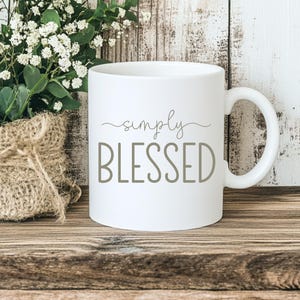 Puede incluir: Una taza de cerámica blanca con asa que presenta el texto simply BLESSED impreso en una fuente gris sans-serif y cursiva. La taza está colocada junto a un pequeño recipiente envuelto en arpillera lleno de follaje verde y flores blancas.