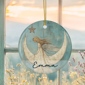 May include: A round, blue ornament with a celestial design. It features an angel seated on a crescent moon, with a star above. The name "Emma" is written in cursive. A gold string is attached for hanging.