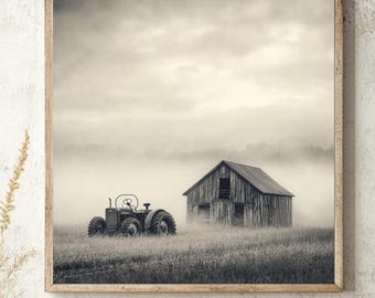 May include: A square wall art print featuring a black and white photograph of a weathered wooden barn and an old tractor sitting in a grassy field. The scene is enveloped in a thick, low-hanging morning fog under a cloudy, overcast sky.