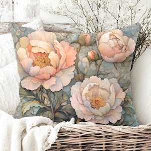May include: Square decorative pillow featuring a watercolor floral design. The pillow showcases large, light pink peonies with yellow centers and green leaves against a muted blue background. The pillow is placed in a wicker basket.