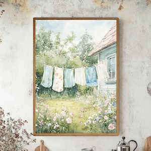 Puede incluir: Pintura a la acuarela de ropa tendida en un tendedero en un jardín soleado. La obra presenta vestidos de colores pastel y una casa azul claro con tejado rojo. La escena está enmarcada en un marco de madera, evocando nostalgia y tranquilidad.