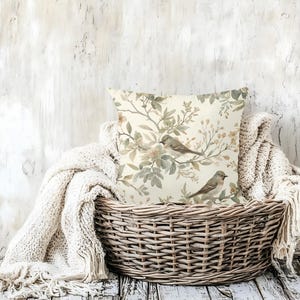 May include: Square cushion with a bird and floral design. The cushion features a cream background with watercolour-style illustrations of birds, leaves, and flowers in shades of green, brown, and beige. The cushion is in a woven basket with a cream-coloured blanket.