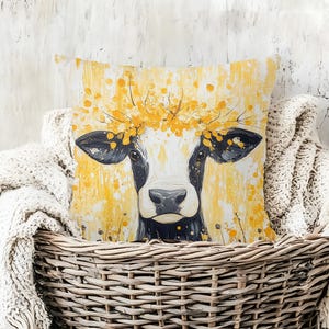 May include: Square decorative pillow featuring a cow with a black and white face, adorned with a yellow floral crown. The background is a textured yellow with paint splatter effects. The pillow is placed in a woven basket.