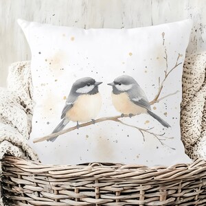 May include: White square throw pillow featuring a watercolor illustration of two small birds perched on a branch. The birds have gray and tan feathers, with a light beige background and gold paint splatters. The pillow is placed in a wicker basket.