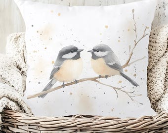 May include: White square throw pillow featuring a watercolor illustration of two small birds perched on a branch. The birds have gray and tan feathers, with a light beige background and gold paint splatters. The pillow is placed in a wicker basket.
