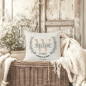 May include: A square, off-white decorative pillow with a family name design. The design includes a large letter "W", the family name "WILLIAMS", and the words "FAMILY welcome to our home" with the year "EST. 2026". The design is surrounded by a floral wreath.