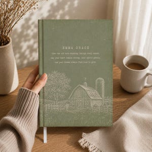May include: A sage green hardcover journal featuring a white line-art illustration of a barn, a silo, and a tree. The text reads: EMMA GRACE. Like the old barn standing through every season, may your heart remain strong, your spirit gentle, and your dreams always find room to grow.