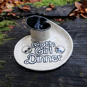 Könnte beinhalten: Beige Keramik-Servierschale mit schwarzer Schüssel und geschwungener Platte. Die Platte trägt den Text "Goth Girl Dinner" und Totenkopf-Illustrationen in Schwarz. Die Schale steht auf einer dunklen Holzoberfläche.