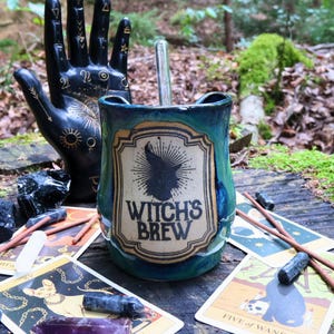 Può includere: Una tazza in ceramica blu e verde acqua con un'etichetta beige recante il testo Witch's Brew e la sagoma di un cappello da strega. La tazza è accompagnata da una scultura a forma di mano nera con simboli celesti dorati e carte dei tarocchi.