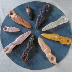 May include: A collection of handmade ceramic sculptures in various colors, including pink, brown, yellow, and blue. The abstract forms are arranged on a blue woven mat. Each piece has a unique shape with a hole or opening.