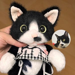 Puede incluir: Un gato de peluche hecho a mano con pelaje blanco y negro, ojos grandes y amarillos y una nariz rosada. El gato lleva un vestido a cuadros rosa y negro con un adorno de flor rosa. Una imagen más pequeña de la cara del gato está en el fondo.