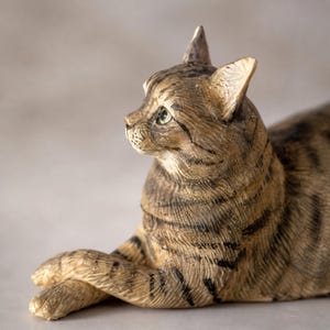 May include: A detailed figurine of a tabby cat in a relaxed pose. The cat has brown and black stripes, green eyes, and is lying down with its paws crossed. The sculpture is made of a material that appears to be ceramic or resin.