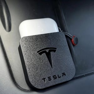 May include: A white wireless earbud charging case housed in a black textured protective cover featuring the embossed Tesla logo and the word Tesla at the bottom. A small black cord with a red ring accent is attached to the side of the case.