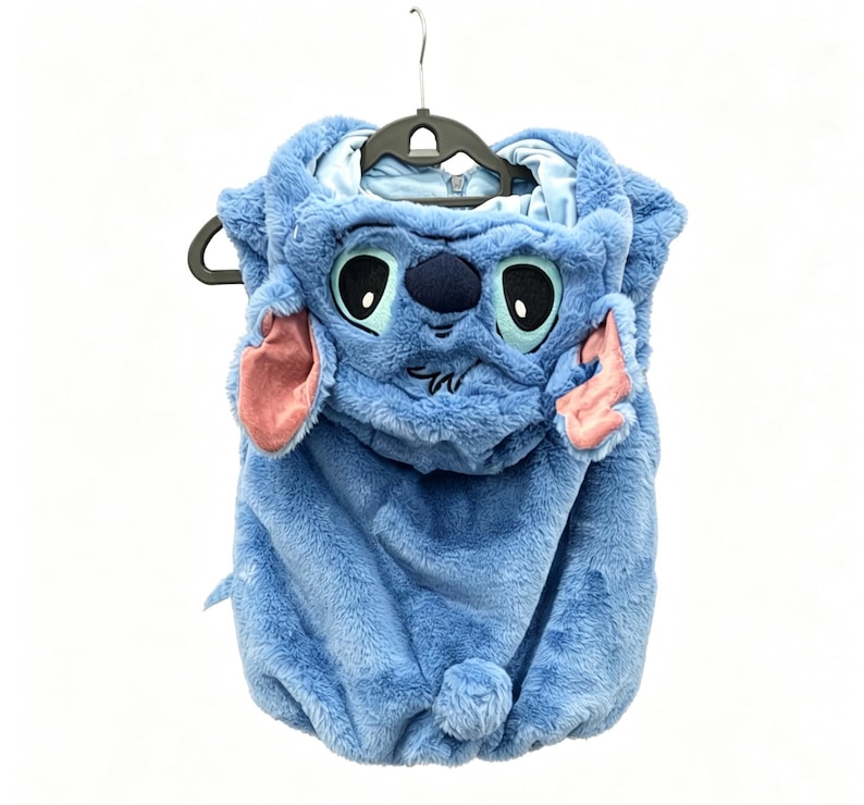 May include: A blue plush costume resembling the character Stitch from the animated film Lilo & Stitch. The costume features a hood with Stitch's face, including large eyes, a dark blue nose, and pink ears. The costume is hanging on a black hanger.