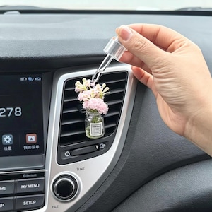 Puede incluir: Un pequeño frasco de vidrio con flores artificiales de color rosa claro sujeto a la rejilla de ventilación de un coche. Un gotero de plástico transparente se sitúa sobre el frasco. El frasco tiene una etiqueta blanca con texto negro.
