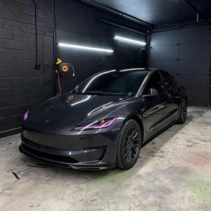 May include: A dark gray sedan with tinted windows and purple accent lighting in the headlights. The vehicle features a sleek, aerodynamic body design with black multi-spoke wheels, parked in a garage with black walls and overhead lighting.