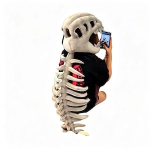 May include: A black t-shirt featuring a large, three-dimensional, off-white skeletal rib cage structure attached to the back, paired with a matching off-white dinosaur skull hood that covers the head, creating a full-body skeletal costume effect.