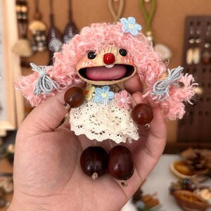 Può includere: Una bambola fatta a mano con capelli di filo rosa, un naso rosso e una bocca spalancata. La bambola ha un vestito di pizzo bianco e tiene due ghiande marroni. Accenti floreali blu e rosa adornano la testa e il vestito della bambola.