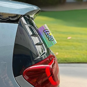 May include: A holographic, cylindrical decal featuring the word LEGO in a bold, black-outlined font with a yellow star accent. The sticker is applied to the rear light assembly of a light blue vehicle, displaying a reflective, iridescent finish.