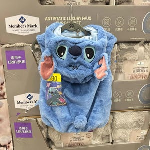 May include: A blue plush costume resembling the character Stitch, featuring large eyes, ears, and a hood. The costume has a tag with the number 30 and the Stitch logo. The product is designed to be a blanket.