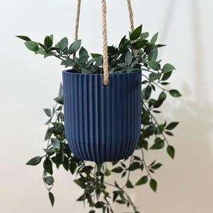 Puede incluir: Una maceta colgante azul marino, acanalada, con una planta verde colgante. La maceta está suspendida por tres cuerdas de cuerda de color natural. Las hojas de la planta caen en cascada desde la maceta.