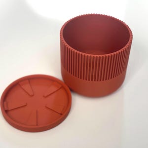 Puede incluir: Una maceta cilíndrica de color terracota con un platillo a juego. La maceta tiene un diseño acanalado en la parte superior y una base lisa. El platillo es redondo con ranuras radiales. Los artículos son de un material mate.