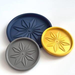 May include: Three round coasters in navy blue, gold, and gray. Each coaster features a raised starburst design in the center. The coasters are made of a solid material and are suitable for various home decor styles.