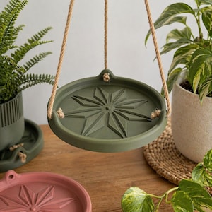 May include: An olive green plant pot holder with a starburst design, suspended by rope. Other plant pots in shades of green and pink are also visible, along with potted plants. The scene is set on a wooden surface, suggesting a home decor theme.