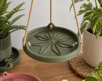 May include: A hanging, olive green plant pot holder with a starburst design, suspended by natural rope. Other plant pots in shades of green and pink are also visible, along with potted plants. The scene is set on a wooden surface, suggesting a home decor theme.