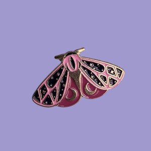 Könnte beinhalten: Eine rosa und schwarze Emaille-Anstecknadel in Form einer Motte mit goldfarbenen Akzenten. Die Anstecknadel hat ein detailliertes Design mit einem Halbmond- und Sternenmuster auf den Flügeln, vor einem hellvioletten Hintergrund.