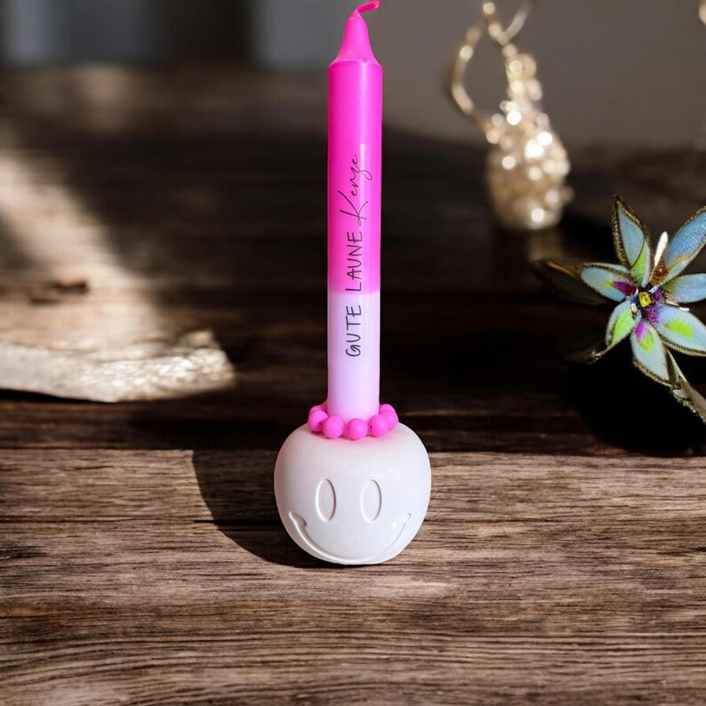 May include: A pink candle with the text "Gute Laune Kerze" is set in a white smiley face candle holder. The candle has a pink top and a white base. The candle holder has two oval eyes and a curved smile.