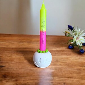 May include: A neon green and pink candle with the text "Gute Laune" is displayed in a white smiley face candle holder. The candle is decorated with green beads at the base. The candle holder is on a wooden surface.
