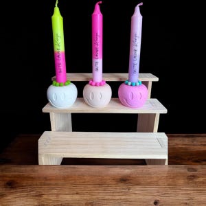 May include: Three colorful candles with smiley face holders. The candles are neon green, pink, and lavender, each with the text "Gute Laune Kerze". The holders are white, peach, and pink, and sit on a small wooden display.