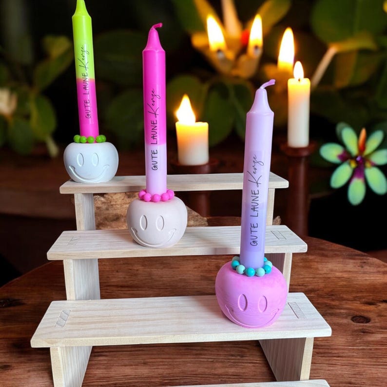 May include: Three colorful candles with the text "Gute Laune Kerze" are displayed on a wooden stand. The candles are green, pink, and purple, each set in a white smiley face holder. Additional lit candles are in the background.