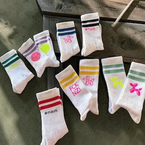 Könnte beinhalten: Eine Kollektion weißer Crew-Socken mit gerippten Bündchen und farbigen Streifen. Die Designs umfassen neonfarbene Smileys, Ballon-Tier-Grafiken sowie Schriftzüge wie #fuck, FCK NZI und #mausi in Farben wie Pink, Gelb und Grün.