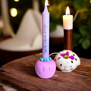 May include: A lit purple candle with the text "GUTE LAUNE KENZE" sits in a pink smiley face candle holder. A white and brown candle and a decorative pumpkin candle holder are also visible on a wooden surface.