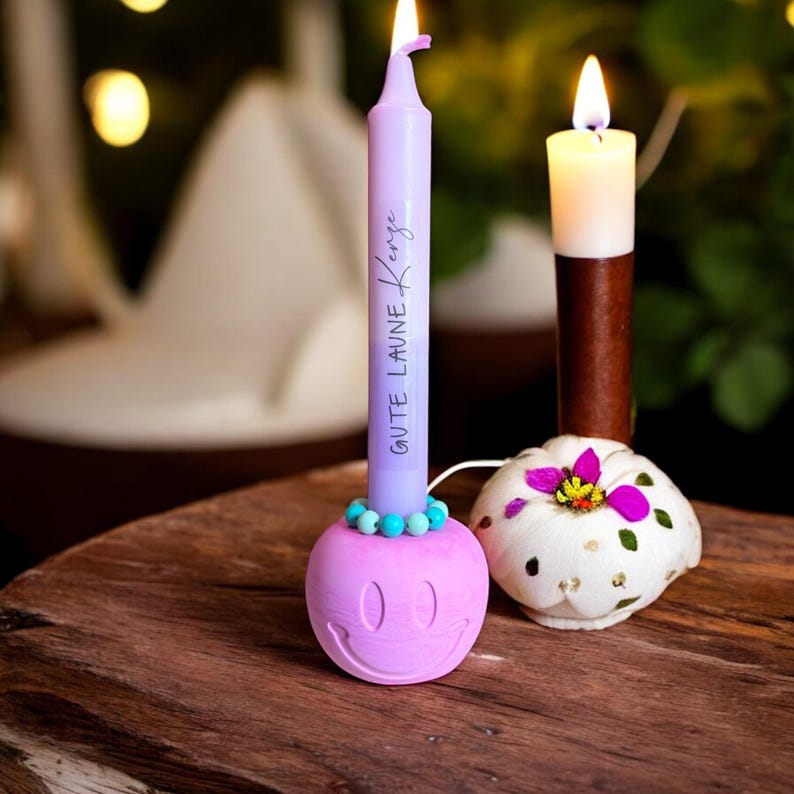May include: A lit purple candle with the text "GUTE LAUNE KENZE" sits in a pink smiley face candle holder. A white and brown candle and a decorative pumpkin candle holder are also visible on a wooden surface.